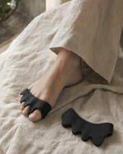 KinetiPath Reflex toe spacers in black worn between the toes, a bare foot resting on linen