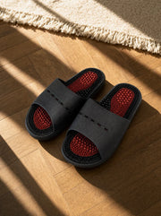 Meridian men's reflexology slides in black with a red footbed, on a warm oak floor beside a linen rug