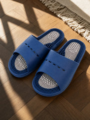 Meridian men's reflexology slides in navy with a pearl grey footbed, on a warm oak floor beside a linen rug